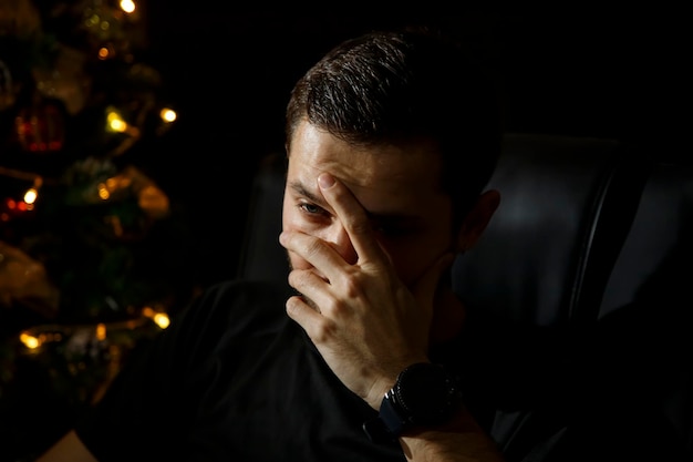 Un hombre cansado, abatido por los problemas que ocultan su rostro con la mano, se sienta junto al árbol de Navidad