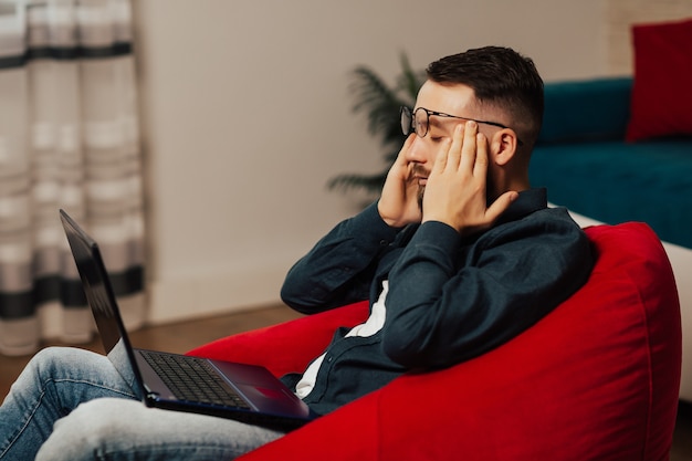 Hombre cansado barbudo guapo joven sentado en un sillón y tratando de trabajar