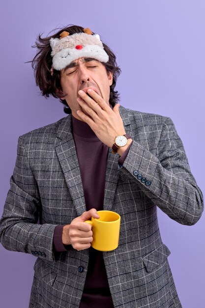 Hombre cansado bostezando sosteniendo una taza amarilla, con antifaz para dormir.