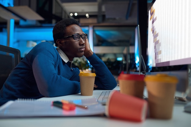 Hombre cansado de gafas trabaja en equipo, estilo de vida de oficina. Persona del sexo masculino en el escritorio, interior oscuro, lugar de trabajo moderno