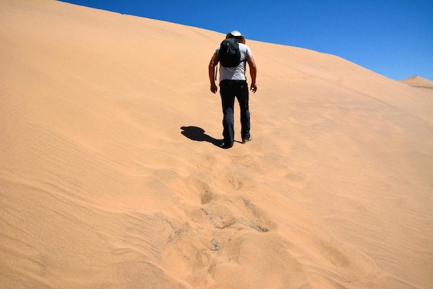 Un hombre cansado con una mochila en la espalda y con una gorra sube una colina de arena del desierto Vista trasera