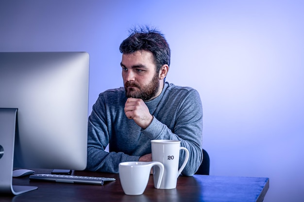 El hombre cansado se sienta frente a una computadora con una taza de café iluminado