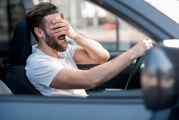 Hombre cansado vestido casual con camiseta blanca conduciendo un coche con dolor de cabeza en la ciudad