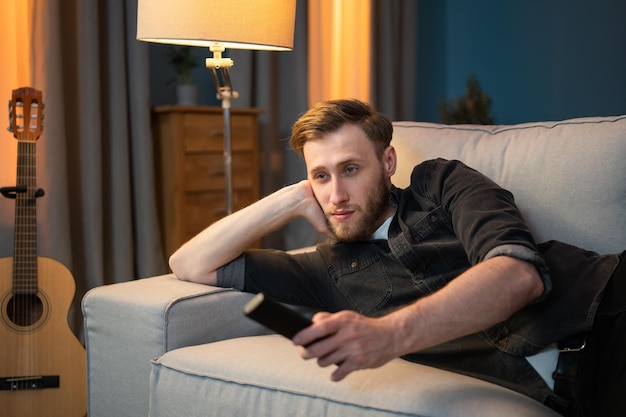 Un hombre cansado yace de lado apoyando la cabeza en el sofá de la sala de estar por la noche con los ojos vidriosos