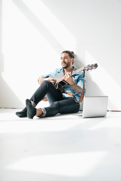 Hombre cantando sentado con una laptop en el piso tocando la guitarra