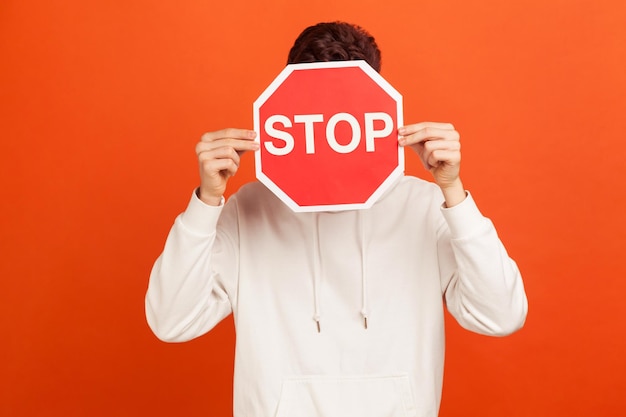 Hombre con capucha blanca casual escondido detrás de la señal de alto, adolescente temeroso de la intimidación, víctima de violencia doméstica. Disparo de estudio interior aislado sobre fondo naranja