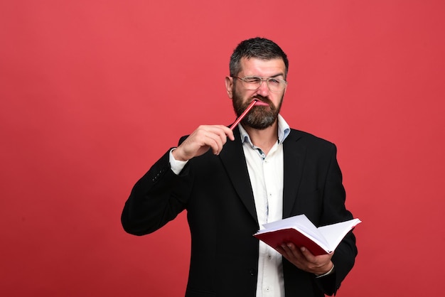 Hombre con cara graciosa sostiene libro y lápiz en la boca sobre fondo rojo.