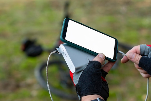 El hombre carga un teléfono inteligente con un banco de energía en el fondo de una bicicleta en el bosque