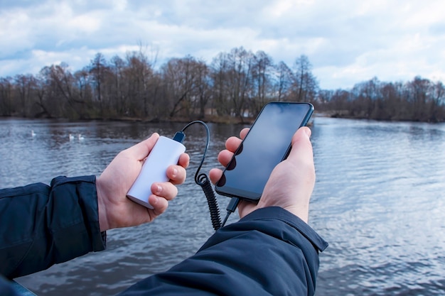 Un hombre carga un teléfono inteligente con un banco de energía. El teléfono en la mano se está cargando con un cargador portátil contra el fondo del lago.