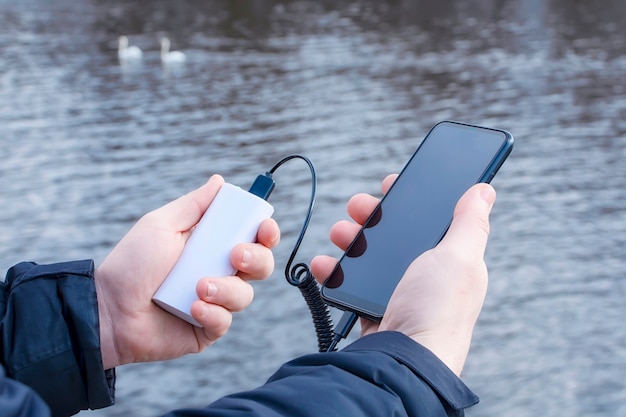 Un hombre carga un teléfono inteligente con un banco de energía. El teléfono en la mano se está cargando con un cargador portátil contra el fondo del lago.