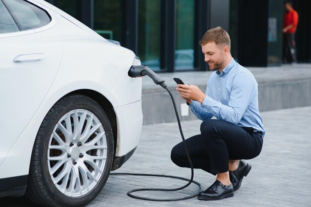 Hombre cargando su coche eléctrico en la estación de carga y usando smartphone