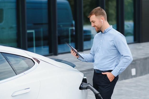 Hombre cargando su coche eléctrico en la estación de carga y usando smartphone