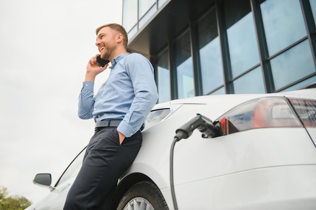 Hombre cargando su coche eléctrico en la estación de carga y usando smartphone