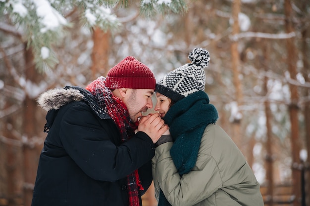 Hombre cariñoso calentando las manos de su mujer con su aliento. Pasan tiempo en un parque en invierno. Ambos con sombreros, chaquetas y bufandas.