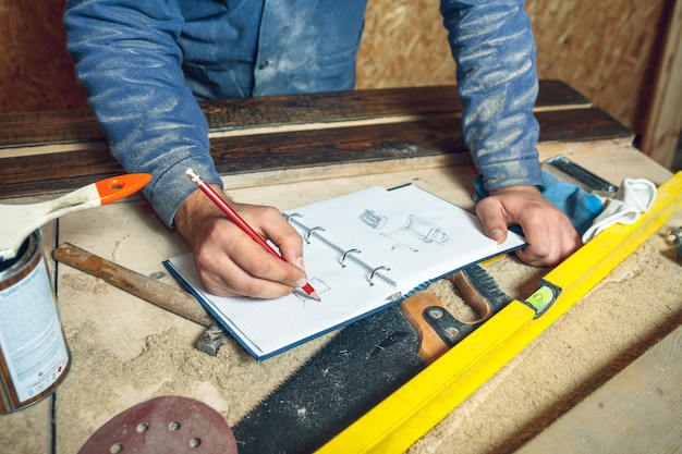 Hombre carpintero en su estudio de casa trabajando con madera y dibuja bocetos a lápiz en un cuaderno