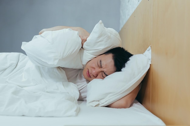 Un hombre en casa intenta quedarse dormido. Los vecinos ruidosos interfieren con el sueño. Un asiático cierra los oídos con almohadas cansados después del trabajo.