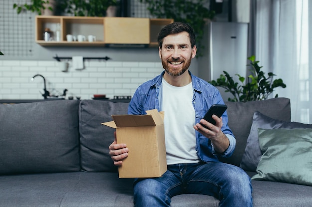 El hombre en casa recibió un pedido de la tienda en línea feliz abre una caja de cartón con un paquete deja una respuesta positiva por teléfono