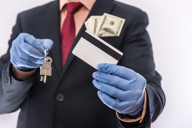 El hombre de la casa de la venta o del alquiler de la seguridad en los guantes médicos tiene la llave de la casa y el dólar. 2019-nCoV