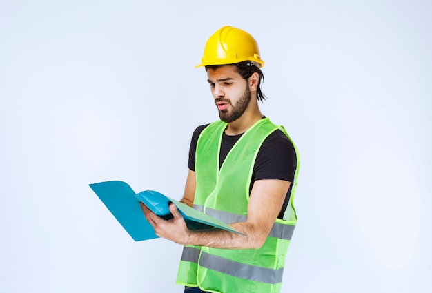 Hombre con casco amarillo sosteniendo y comprobando la carpeta del proyecto azul.