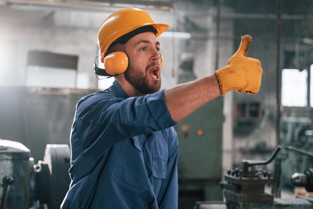 Foto hombre con casco y auriculares está mostrando el pulgar hacia arriba trabajador de fábrica en uniforme azul está en el interior