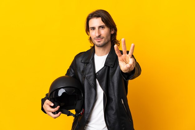 Hombre con un casco de moto aislado sobre fondo amarillo feliz y contando tres con los dedos