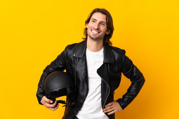 Hombre con un casco de motocicleta aislado sobre fondo amarillo posando con los brazos en la cadera y sonriendo