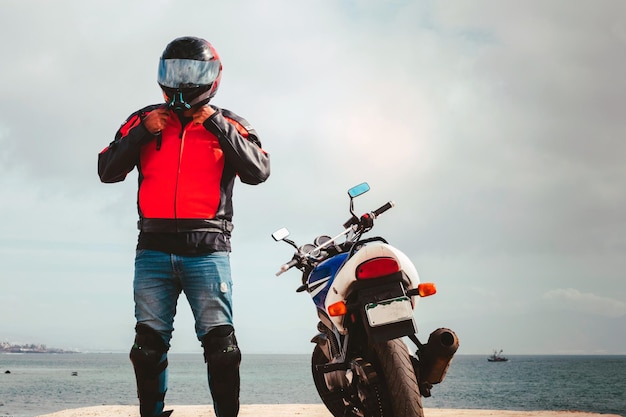 Foto un hombre con casco de motocicleta se para al lado de su motocicleta en una playa.