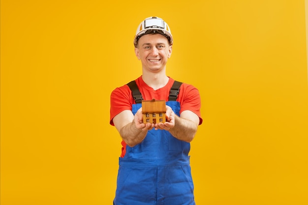 Hombre con casco de seguridad y uniforme de trabajo sostiene una pequeña casa de juguete Mantenimiento de casas Construcción inmobiliaria