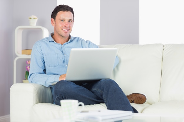 Hombre casual contenido que se sienta en el sofá usando la computadora portátil que mira la cámara