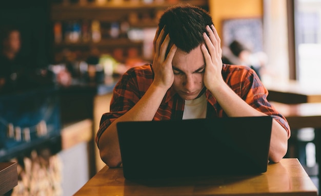 Hombre casual feliz con teléfono inteligente y computadora portátil en café bajo estrés y preparándose para exámenes