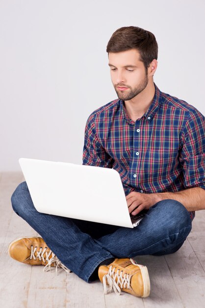 Hombre casual que trabaja en la computadora portátil. Apuesto joven trabajando en la computadora portátil mientras está sentado en el piso de madera