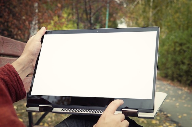 Foto hombre casual sentado en un banco en el parque de otoño con las piernas cruzadas y presentando su computadora portátil con pantalla blanca en blanco pantalla táctil en la computadora portátil