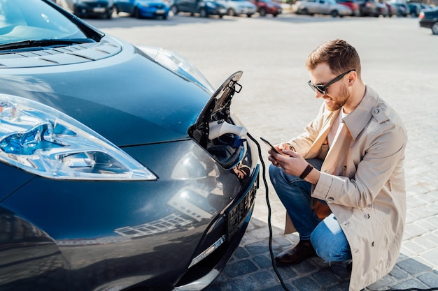 Hombre casual con smartphone cerca de coche eléctrico