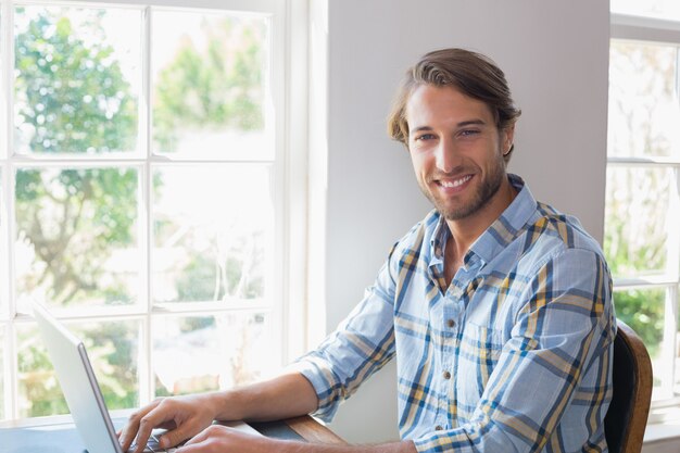 Hombre casual sonriente que usa el ordenador portátil que mira la cámara