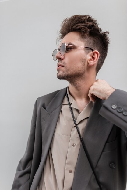 Hombre casual urbano hipster modelo masculino con gafas de sol, camisa y chaqueta de pie cerca de un fondo gris.