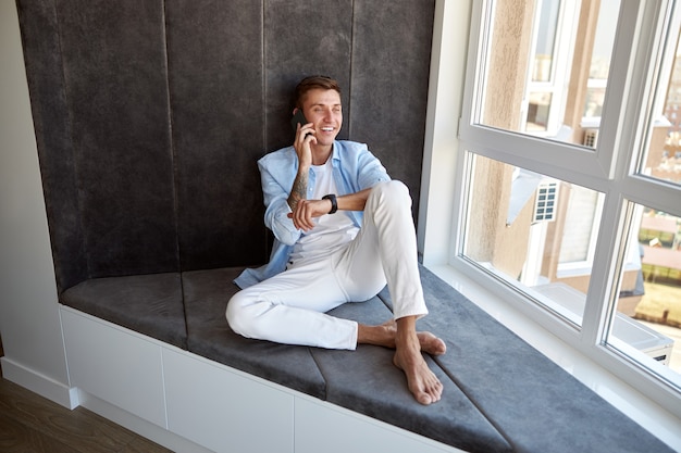 Hombre caucásico adulto feliz está sentado cerca de la ventana panorámica y se enfría