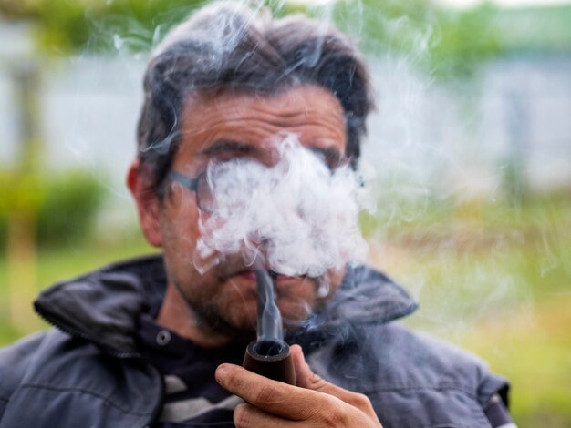 Un hombre caucásico sin afeitar con gafas fuma una pipa.