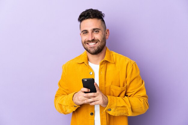 Hombre caucásico aislado en la pared púrpura mirando a la cámara y sonriendo mientras usa el móvil