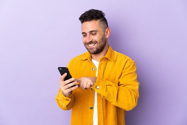 Foto hombre caucásico aislado sobre fondo morado enviando un mensaje o correo electrónico con el móvil