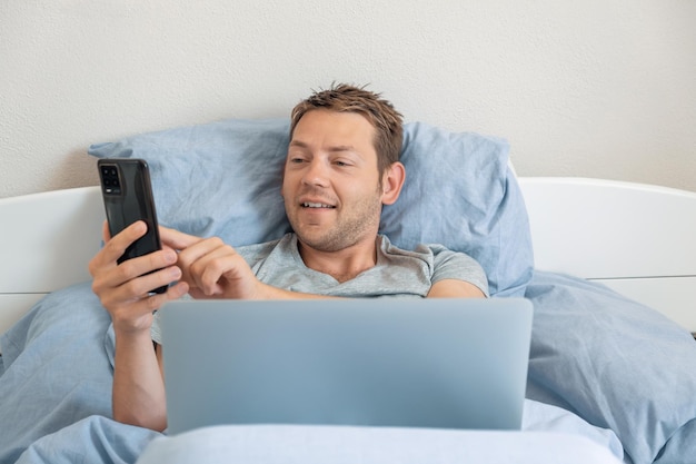 Hombre caucásico alegre sonriendo y enviando mensajes de texto en el teléfono inteligente durante el chat de video en el portátil masculino