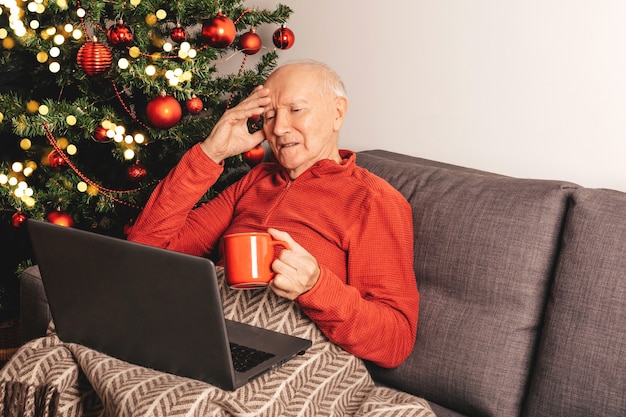 Hombre caucásico anciano solitario triste con portátil sentado en un sofá cerca de un árbol de Navidad con taza de té charlando con familiares en línea. Autoaislamiento, estado de ánimo de vacaciones.