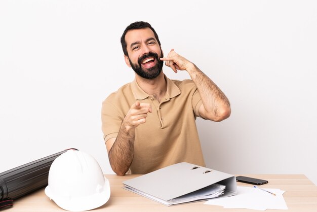 Hombre caucásico arquitecto con barba en una mesa haciendo gesto de teléfono y apuntando hacia el frente.