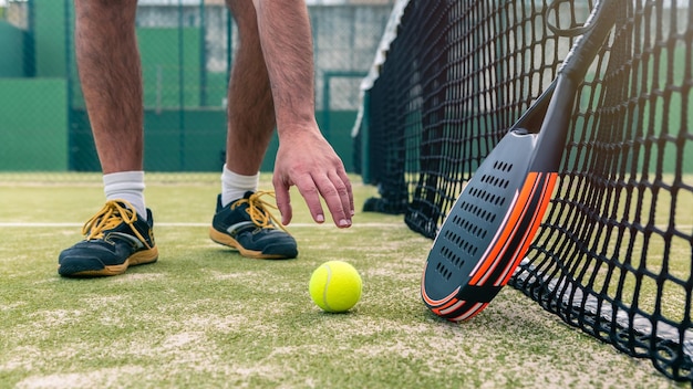 Un hombre caucásico atrapa una pelota amarilla cerca de la raqueta de pádel en la hierba verde de la cancha