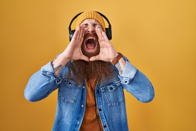 Hombre caucásico con barba larga escuchando música usando auriculares gritando enojado con las manos sobre la boca