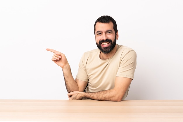 Hombre caucásico con barba en una mesa apuntando con el dedo hacia un lado.