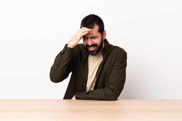 Hombre caucásico con barba en una mesa con dolor de cabeza.