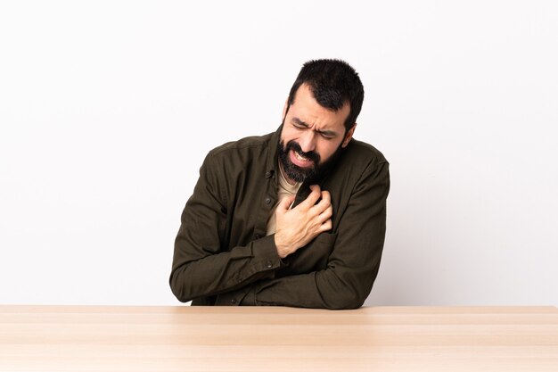 Hombre caucásico con barba en una mesa con dolor en el corazón.