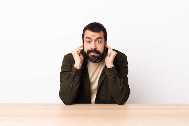 Hombre caucásico con barba en una mesa frustrado y cubriendo las orejas