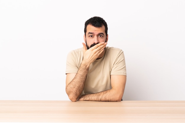 Hombre caucásico con barba en una mesa que cubre la boca con la mano.