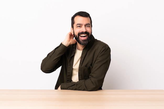 Hombre caucásico con barba en una mesa riendo.